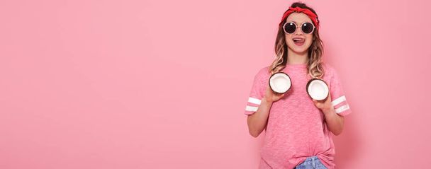 Beautiful young girl in pink t-shirt and glasses holding funny sexy coconuts on pink background - Fotografie, Obrázek