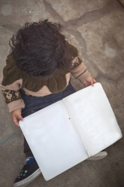 Μικρό παιδί κορίτσι κρατά βιβλίο, κάθεται στο πάτωμα και κοιτώντας ψηλά στο camera - Φωτογραφία, εικόνα