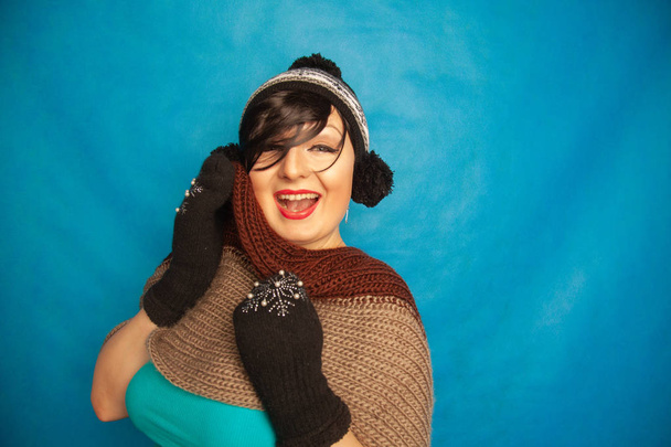 pretty brunette girl wearing fashion black knitted winter hat and warm beautiful mittens on blue studio solid background - Fotoğraf, Görsel