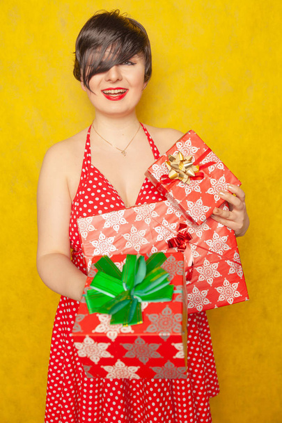 γλυκό χαρούμενο κορίτσι σε ένα φόρεμα κόκκινο πουά ρετρό στέκεται με τρία κουτιά δώρων με τα χέρια και τα χαμόγελα ειλικρινά σε κίτρινο στερεό φόντο στο στούντιο - Φωτογραφία, εικόνα