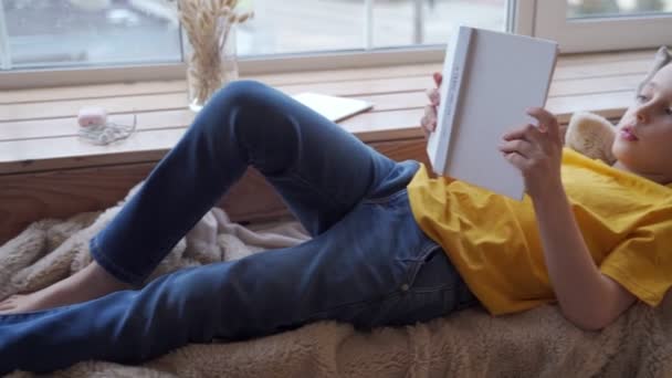 Cute little boy lying on windowsill and reading book - Séquence, vidéo