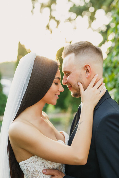 young beautiful wedding couple bride and groom are standing in the park at sunset and smiling bride in beautiful dress groom is dressed stylishly - 写真・画像