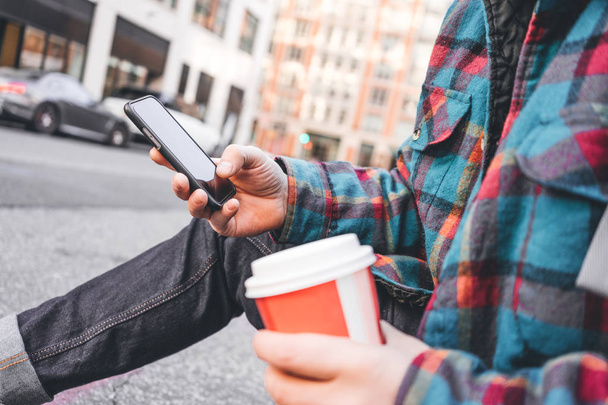 Hipster man rust met een kop warme koffie op de stoep en texting massage stad op mobiele telefoon scherm. Man met behulp van smartphone en zittend op stedelijke stoep weg - Foto, afbeelding