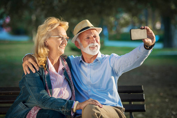 Senior couple taking a selfie photo with smart phone in a park - Фото, изображение