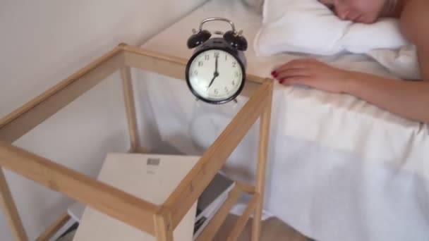 Young beautiful woman hates waking up early in the morning. Sleepy girl looking at alarm clock and trying to hide under the pillow - Imágenes, Vídeo