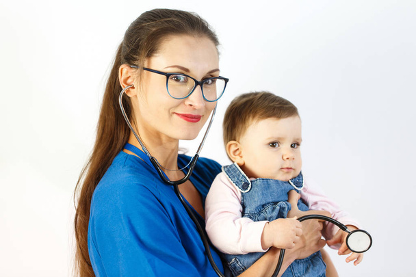 Egy fiatal nő orvos a baba tartja a karjában, ő mosolyog, és a gyermek játszik a sztetoszkóp. - Fotó, kép