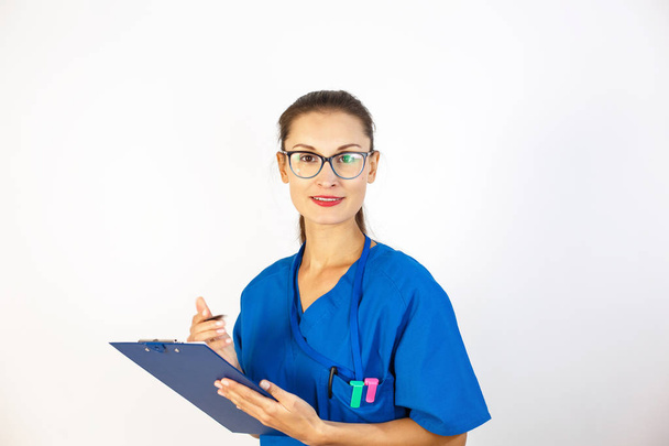 Eine Ärztin im blauen Gewand schreibt Daten an ein Tagebuch, sie trägt ein Stethoskop um den Hals, sie blickt in die Kamera. weißer Hintergrund - Foto, Bild