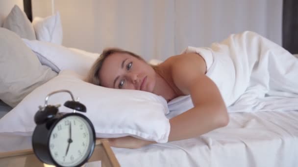 Young beautiful woman hates waking up early in the morning. Sleepy girl looking at alarm clock and trying to hide under the pillow - Video, Çekim