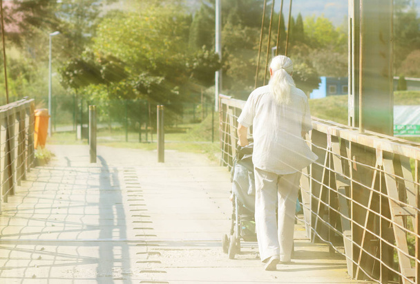 Un anciano con pelo largo, gris y atlético camina con un niño en un cochecito sobre un puente. Actividad en la vejez. Lecciones para que los abuelos cuiden a sus hijos. Jubilosa vejez con nietos
 - Foto, imagen