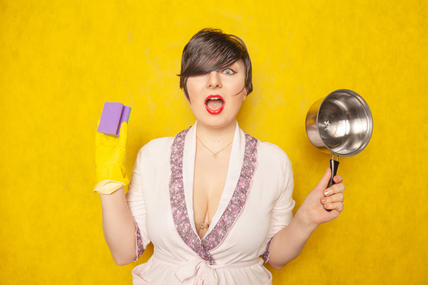 красивая светлая брюнетка в халате держит сковороду и губку для мытья посуды. молодая женщина домохозяйка на жёлтом фоне студии
.  - Фото, изображение