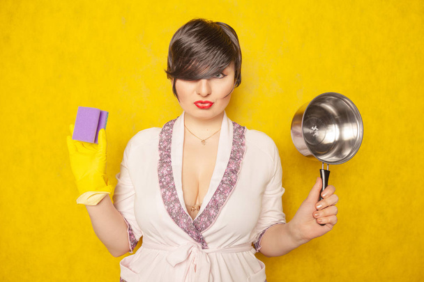 beautiful bright brunette in a Bathrobe holds a pan and a sponge for washing dishes. young woman housewife on yellow studio background.  - Foto, Bild