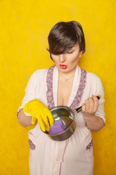 beautiful bright brunette in a Bathrobe holds a pan and a sponge for washing dishes. young woman housewife on yellow studio background.  - Foto, Imagem