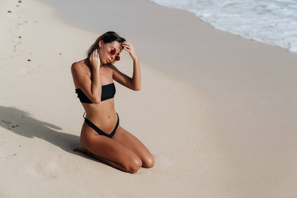 Sexy tanned girl in black swimsuit and sunglasses posing on sandy beach - Фото, изображение