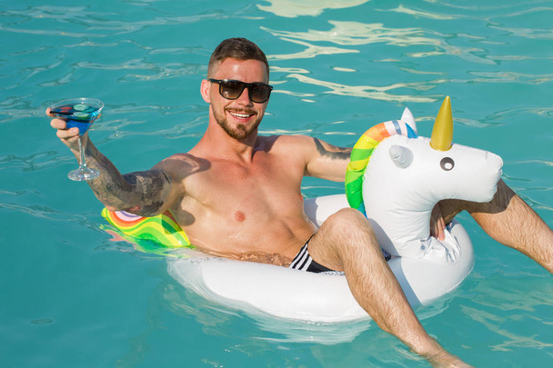 Cheerful handsome bearded man cheering with his cocktail glass while swimming on inflatable unicorn at the swimming pool. Happy man relaxing during his summer vacation. Lifestyle, partying, travel concept - Photo, Image