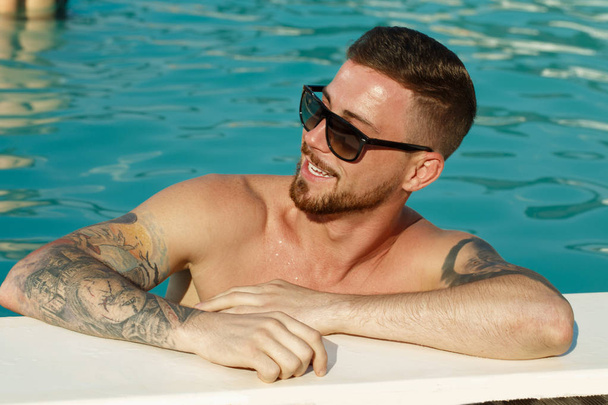 Közelről portré egy szép szakállas férfi napszemüveg mosolyogva nézett el a medence mellett pihenve. Boldog ember pihen úszás után. Nyári, utazás, szabadidő, szabadidő-koncepció - Fotó, kép