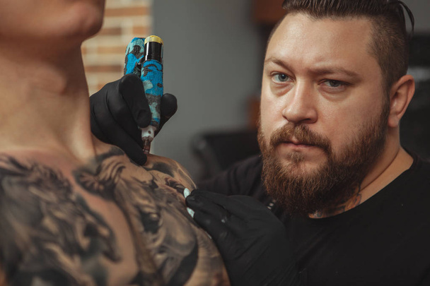 Бородатый татуировщик делает татуировки на теле своего клиента
 - Фото, изображение