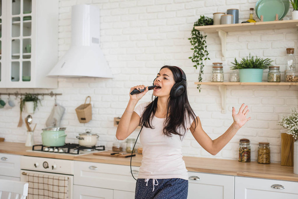 Young happy woman  in headphones dancing and singing in kitchen and having some fun - Фото, изображение