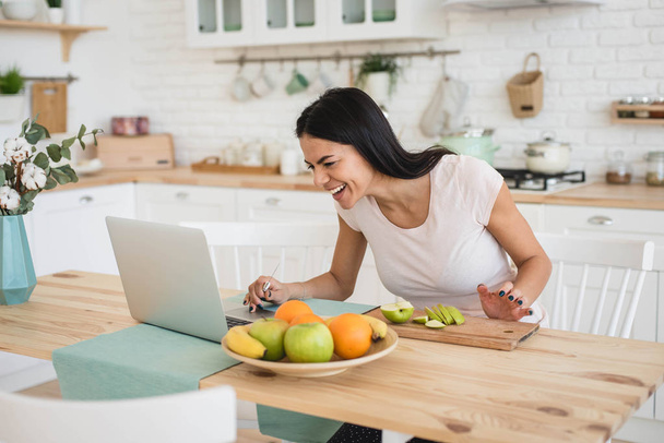 Young attractive woman using laptop in kitchen while cooking - Foto, Imagen