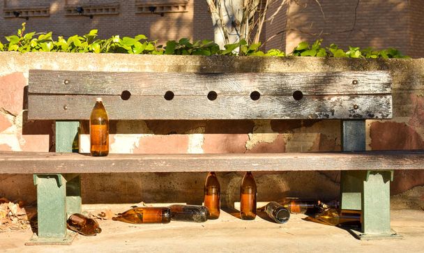 πολλά μεγάλα πορτοκαλί μπουκάλια μπύρας, κατασκευασμένο από γυαλί εντελώς άδειο στο πάρκο λόγω κάποιος έχει πιει χρόνο πριν από την αναχώρηση τους πάνω και κάτω από έναν πάγκο καφέ σε μια ηλιόλουστη μέρα - Φωτογραφία, εικόνα