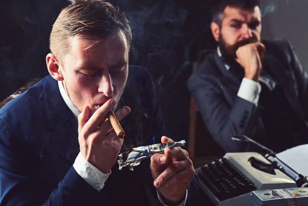 He is lousy with money. Rich man smoking during business meeting. Man light up cigar from money banknote. Businessmen have money to burn. Business partners writing financial report. Waste of money - Фото, зображення