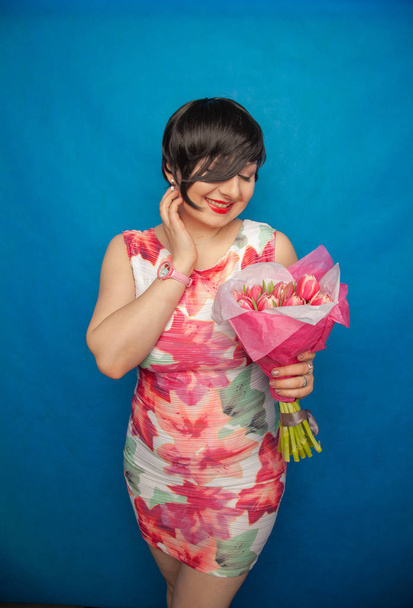 Nettes lächelndes Mädchen im Frühlingskleid hält im Atelier einen rosafarbenen, frischen Strauß Tulpenblumen auf blauem Hintergrund. Sie genießt das Geschenk wirklich und ist absolut glücklich. - Foto, Bild