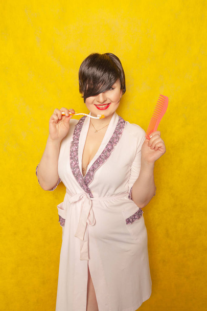 cute chubby girl in a pink robe stands with a comb and toothbrush on a yellow background in the Studio - Photo, Image