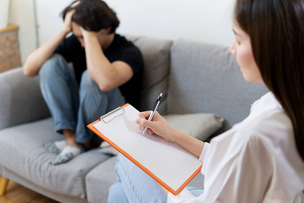 Young male patient sitting on sofa with sad face and hands on head is having consultant by professional psychologist female. Psychological consultant concept. - Photo, Image