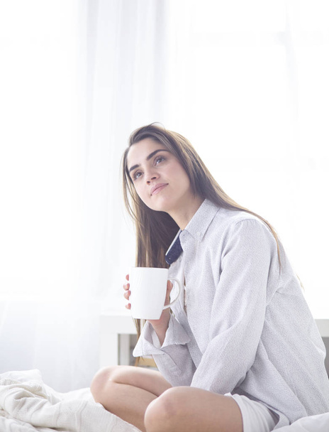 Молодая красивая счастливая брюнетка в черном шелковом халате лежит в постели, пьет утренний кофе, белая чашка. Женский день
 - Фото, изображение
