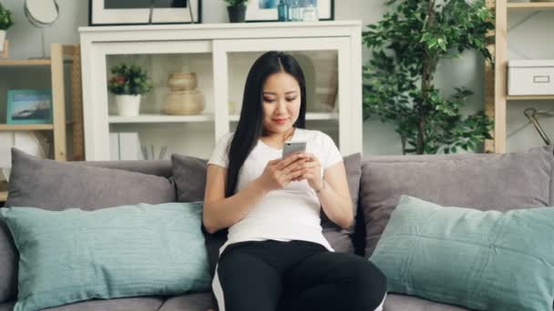 Cute Asian girl is using smartphone touching screen and smiling relaxing on sofa at home alone enjoying modern technology. Youth and gadgets concept. - Felvétel, videó