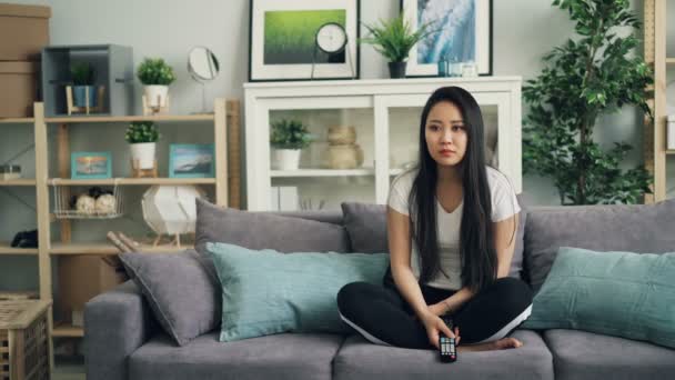 Portrait of good-looking Asian girl using remote control sitting on sofa at home and watching TV. Young woman in casual clothing is smiling and laughing. - Séquence, vidéo