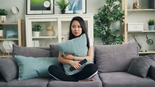 Beautiful Asian young woman is watching horror film on TV at home holding pillow and reacting to scary episodes then using remote control. Movies and people concept. - Кадри, відео