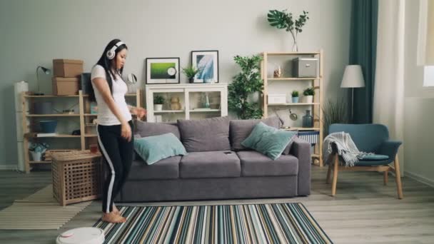 Asian young lady is enjoyig modern technology listening to music in wireless headphones and dancing while robotic hoover is cleaning floor in her nice apartment. - Séquence, vidéo