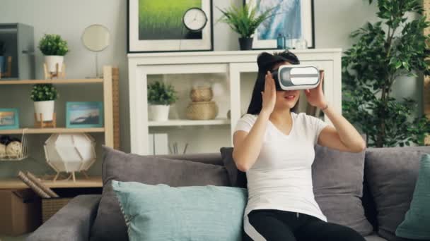 Beautiful dark-haired brunette is having fun with virtual reality glasses moving head and hands wearing headset. Girl is sitting on sofa in nice modern house. - Metraje, vídeo
