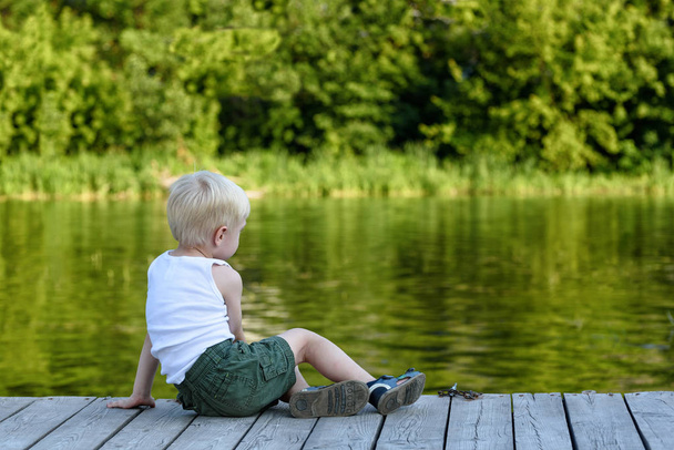 Little blond boy sitting on a pier on the bank of the river amid a green forest - Foto, imagen