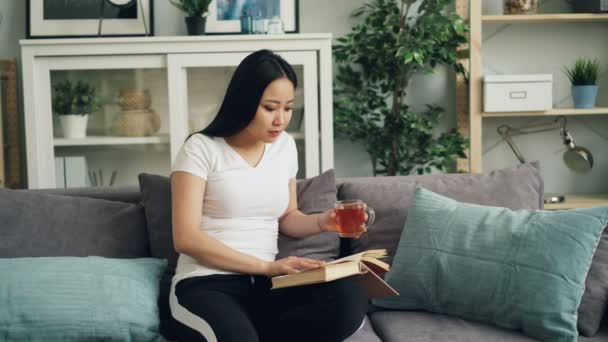 Beautiful Asian girl is reading book and drinking tea sitting on sofa at home and enjoying free time and peaceful occupation. Women, hobby and house concept. - Filmmaterial, Video