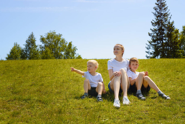 Mutter und zwei kleine Söhne sitzen auf Gras vor einem Hintergrund aus Bäumen und blauem Himmel - Foto, Bild