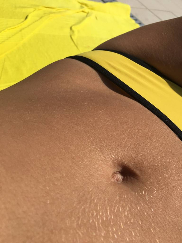 Part of a body of the suntanned girl with a stomach and a navel in a yellow bathing suit, a beach photo of suntanned skin - Photo, Image