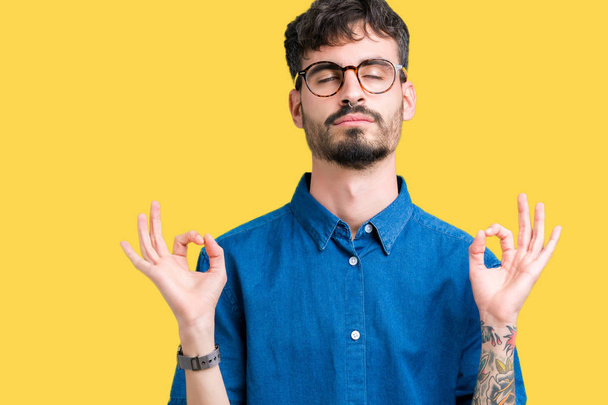 Young handsome man wearing glasses over isolated background relax and smiling with eyes closed doing meditation gesture with fingers. Yoga concept. - Photo, Image