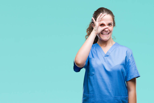 junge brünette Ärztin trägt Krankenschwester oder Chirurg Uniform über isoliertem Hintergrund tun ok Geste mit der Hand lächelnd, Auge durch Finger mit glücklichem Gesicht. - Foto, Bild
