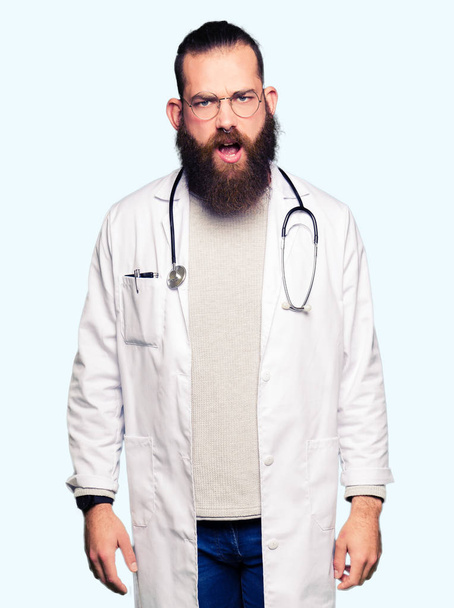 Young blond doctor man with beard wearing medical coat In shock face, looking skeptical and sarcastic, surprised with open mouth - Photo, Image