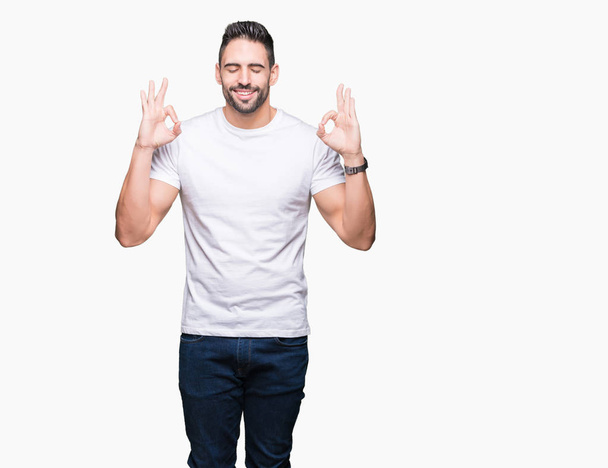 Young man wearing casual white t-shirt over isolated background relax and smiling with eyes closed doing meditation gesture with fingers. Yoga concept. - 写真・画像