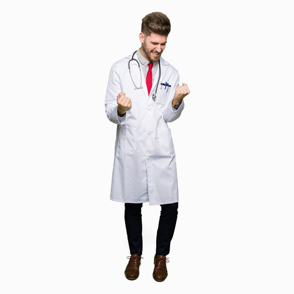 Young handsome doctor man wearing medical coat very happy and excited doing winner gesture with arms raised, smiling and screaming for success. Celebration concept. - Photo, image