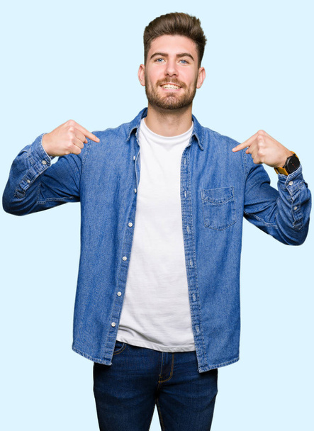 junger gutaussehender blonder Mann in lässiger Jeansjacke, der selbstbewusst mit einem Lächeln auf dem Gesicht aussieht und stolz und glücklich mit den Fingern zeigt. - Foto, Bild