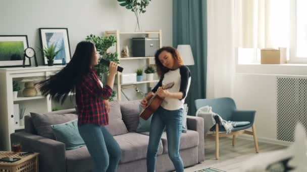 Joyful young women African American and Asian in casual clothing are playing the guitar, dancing and singing in TV remote control having fun in modern apartment. - Кадры, видео