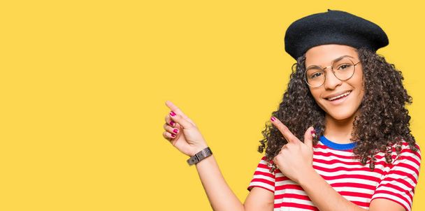 Young beautiful woman with curly hair wearing glasses and fashion beret smiling and looking at the camera pointing with two hands and fingers to the side. - Фото, зображення