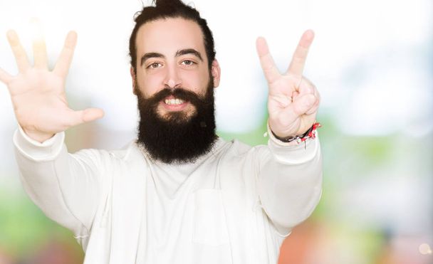 Молодой человек с длинными волосами и бородой в спортивной толстовке показывает и показывает пальцами номер семь улыбаясь уверенно и счастливо
. - Фото, изображение