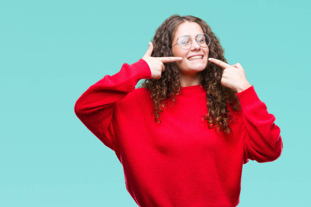 Красива брюнетка кучеряве волосся молода дівчина в окулярах і зимовий светр над ізольованим тлом посміхається впевнено показуючи і вказуючи пальцями зуби і рот. Концепція здоров'я
. - Фото, зображення