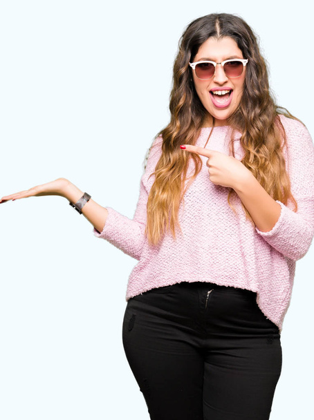 Giovane bella donna indossa occhiali da sole e maglione rosa stupito e sorridente alla fotocamera mentre si presenta con la mano e punta con il dito
. - Foto, immagini