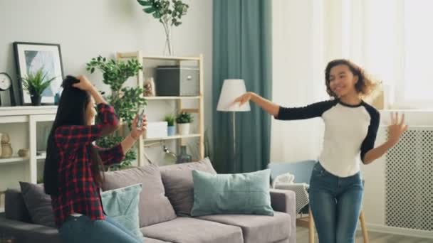 Emotional African American girl student is dancing at home laughing and having fun while her Asian friend is recording video with smartphone camera. Youth and technology concept. - Metraje, vídeo