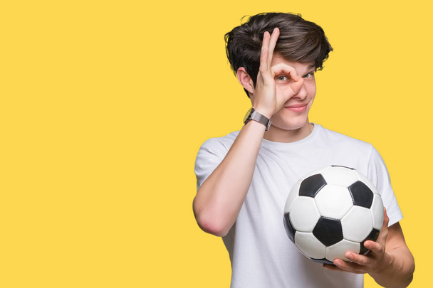Молодий чоловік тримає футбольний м'яч над ізольованим тлом з щасливим обличчям, посміхаючись, роблячи знак ОК з рукою на око дивиться крізь пальці
 - Фото, зображення
